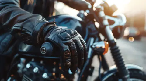 A person's hand wearing a leather motorcycle glove on the handlebars of a motorcycle.
